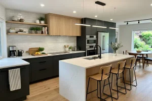Finished kitchen remodel with two-tone cabinetry in charcoal and natural wood, white marble-look countertops, a spacious island with bar seating, modern black fixtures, stainless steel appliances, and warm wood flooring.